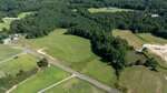Aerial view of a rural landscape featuring open grassy fields, a two-lane road, dense woods, a small pond, and scattered houses surrounded by trees under clear skies.