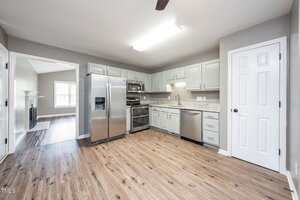 Modern kitchen with stainless steel appliances, light gray cabinets, granite countertops, wood-style flooring, and neutral-colored walls. The space connects to an adjacent room with a window and fireplace.