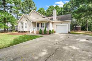 Single-story beige house with a front porch, attached one-car garage, and a wide driveway. The yard has grass and mulch with small shrubs and is surrounded by tall pine trees. A wooden shed is visible in the background.