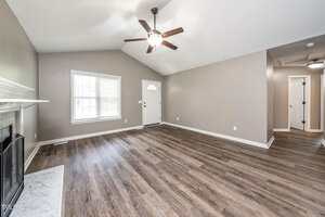 A spacious living room with light brown wood-look flooring, beige walls, a white front door, a white-framed window, a ceiling fan with light, and a fireplace with a white mantle on the left.