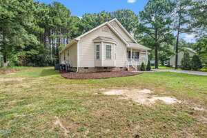 A single-story beige house with white trim, a small front porch, and brick foundation, surrounded by a grassy yard, trees, and a driveway on the right side.