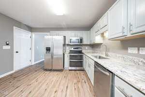 Modern kitchen with stainless steel appliances, light gray cabinets, granite countertops, and wood-look flooring. The room is well-lit, and the walls are painted a neutral gray color.