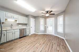 Bright kitchen with light gray cabinets, granite countertops, stainless steel dishwasher, wood-look flooring, a ceiling fan, and double doors leading outside. Two windows provide additional natural light.