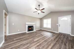 A spacious living room with light gray walls, wood-style flooring, a white front door, a large window, a ceiling fan with a light, and a white fireplace with a mantel. The room is empty and well-lit.