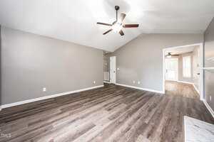 A spacious, empty living room with light gray walls, wood-look vinyl flooring, a ceiling fan with lights, and an open doorway leading to another bright room with similar flooring.