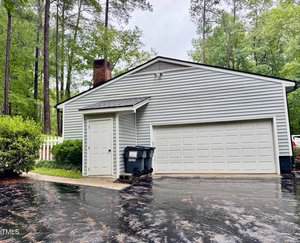 A gray single-story house with white trim, a two-car garage, a small covered side entrance, two trash bins, and a paved driveway surrounded by tall trees and greenery.