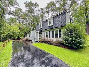 Charming two-story house at 3070 Cameron Drive, Henderson, with dark shingles and white siding. Dormer windows overlook a wet driveway bordered by green grass, tall trees, and a wooden fence along the left side.