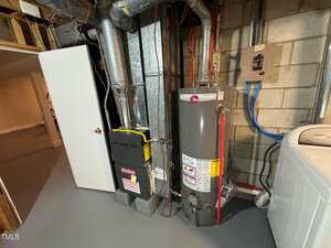 A basement utility area with a water heater, furnace, and exposed ducts next to a concrete wall. A white door is open, and a washing machine is partially visible on the right side.
