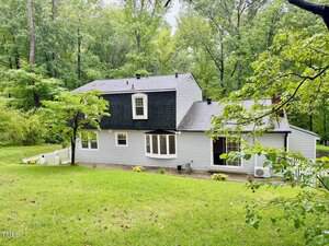 A two-story gray house with white trim, a black roof, and a bay window, surrounded by green grass and trees. A small white picket fence lines part of the property. The setting is lush and wooded.