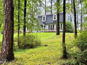 A two-story house with a dark gambrel roof and white siding is surrounded by trees and greenery on a gently sloping lawn. The front porch has a wooden railing and the home sits in a wooded area.