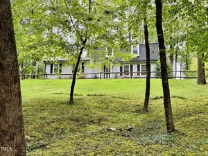 A two-story house with gray siding and white trim is partially visible behind several green trees and a wooden fence, set on a grassy yard.