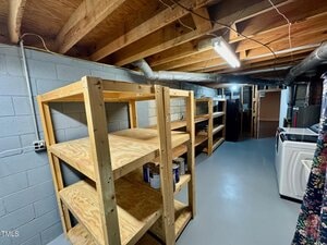 A basement room with exposed wooden ceiling beams, concrete block walls, wooden shelving units, paint cans on shelves, a washer and dryer, ductwork, and fluorescent lighting.