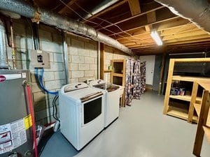 A basement laundry area with a washer and dryer, exposed pipes, water heater, concrete block walls, wooden shelves on the right, a hanging shower curtain, and a closed door at the back.