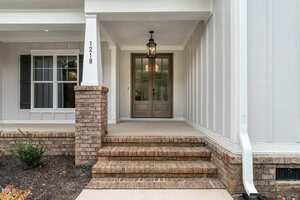 A front porch with three brick steps leading to double wooden doors, a hanging lantern light above, white vertical siding, a brick column, house number 1218, and a white downspout on the right.