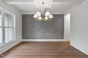 A dining room with a geometric accent wall painted gray, a gold chandelier with white lampshades, large windows on the left, and wood flooring. The room is empty with white walls and trim.