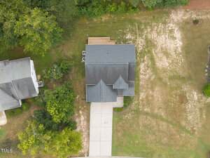 Aerial view of a single-family house with a dark roof, concrete driveway, and a wooden deck at the back. The yard has some grass and bare patches of soil. Neighboring house and trees are visible on the left side.