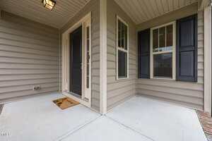 A covered front porch with beige siding, a black front door, a welcome mat, and a window with black shutters. The porch has a concrete floor and a ceiling light.