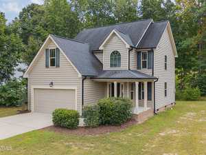 Two-story beige house with black roof, attached two-car garage, covered front porch, and neatly trimmed bushes in front. Surrounded by trees and greenery with a concrete driveway leading to the garage.