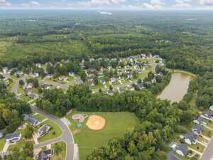 Aerial view of a suburban neighborhood with houses surrounded by trees, a curving road, a baseball field, a playground, and a small pond, bordered by a dense forest under a cloudy sky.