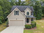 Located at 1976 Ferbow Street in Creedmoor, this two-story beige house features dark shutters, a double garage, a small porch, and neatly trimmed bushes, all set against scenic trees and a lawn with minimal landscaping.
