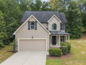 Located at 1976 Ferbow Street in Creedmoor, this two-story beige house features dark shutters, a double garage, a small porch, and neatly trimmed bushes, all set against scenic trees and a lawn with minimal landscaping.