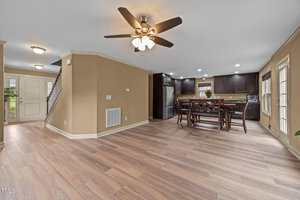 Open-concept living and dining area with wood flooring, ceiling fan, beige walls, staircase by the entrance, and a kitchen with dark cabinets and stainless steel appliances in the background.