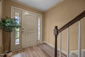 A beige entryway features a front door with two narrow windows on either side, a potted artificial plant on the left, and a staircase with a dark wood handrail and white spindles on the right.