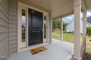 A covered front porch with beige siding and a black door, flanked by windows. A doormat with sunflowers and "welcome" written on it lies in front of the door. The porch overlooks a grassy yard and neighboring homes.