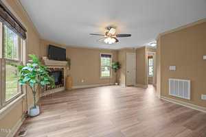 Spacious living room with light wood flooring, tan walls, a ceiling fan with lights, a corner fireplace with a TV, large windows with blinds, and a potted plant. A hallway and doorways are visible in the background.