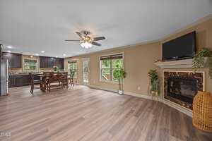 Open-concept living and dining area with wood flooring, ceiling fan, dark kitchen cabinets, large windows, a corner fireplace with a TV, and indoor plants against beige walls.