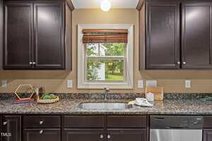 A kitchen countertop with a stainless steel sink below a window, dark wood cabinets, a dishwasher, a basket with decor, a cookbook, a plant, and a towel and soap by the sink. A green yard is visible through the window.
