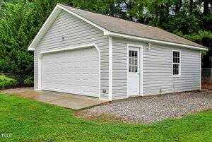 A detached two-car garage with light gray siding, white trim, a white double garage door, and a single white entry door. The structure sits on a concrete and gravel base surrounded by green grass and trees.