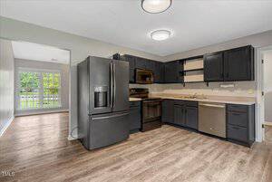 A modern kitchen with dark cabinets, light countertops, stainless steel appliances, and wood-look flooring. An adjacent dining area with large windows and a view of greenery is visible in the background.