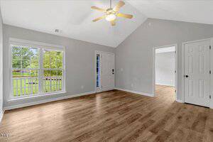 A bright, empty room with light gray walls, a ceiling fan with light, large window, wood flooring, and two white doors leading to other rooms. A view of green trees is visible through the window.