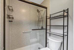 A bathroom with a glass-enclosed shower, a mounted showerhead, two wire shelves inside the shower, a grab bar, a white toilet, and a black metal shelving unit above and beside the toilet.