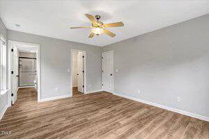 A vacant room with light gray walls, wood-style flooring, a ceiling fan with lights, and three doors leading to a bathroom, closet, and another room. Natural light enters through two windows on the left.