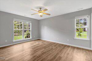 A spacious, empty room with light gray walls, two large windows, a ceiling fan with a light fixture, and wood-look flooring. The windows overlook a green outdoor area.