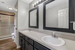 A bathroom with a double sink vanity, two mirrors above the sinks, black cabinets, and a walk-in shower with a glass door. The walls are light gray and the floor has a wood-like finish.