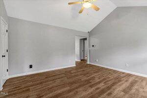 Empty room with light gray walls, wood-style flooring, a ceiling fan with lights, and a white door on the left. The room has a vaulted ceiling and an open doorway leading to another area.