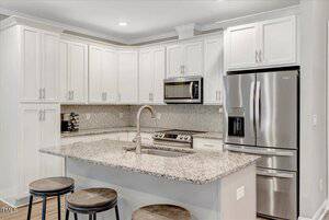 A modern kitchen with white cabinets, stainless steel appliances, a granite countertop island with a sink, three wooden stools, and a herringbone-patterned backsplash.