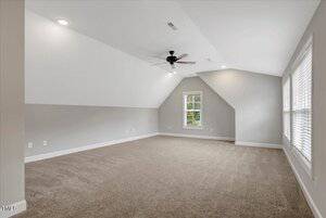 A spacious, empty room with beige carpet, light gray walls, sloped ceiling, ceiling fan, recessed lighting, and two large windows with blinds letting in natural light.
