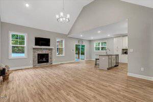 Open-concept living area with wood flooring, a fireplace with stone surround, two windows, and a wall-mounted TV. The space connects to a kitchen with white cabinetry, an island, and stainless steel appliances.