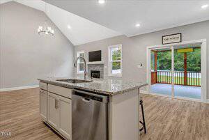 A modern kitchen with a granite island, sink, and dishwasher opens to a living area with wood flooring, light gray walls, multiple windows, and glass doors leading to a screened porch with a wooded view.