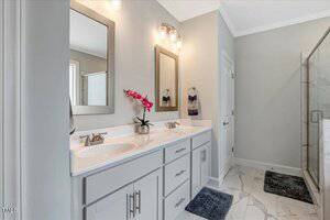 Modern bathroom with a double-sink vanity, two mirrors, wall-mounted lights, a potted orchid, gray cabinets, marble tile floor, two dark rugs, and a glass shower enclosure.