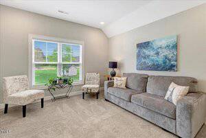 A living room with a large window, two patterned chairs, a small glass table, a gray sofa with pillows, a blue table lamp, and a blue abstract painting on a beige wall. The room has carpeted flooring.