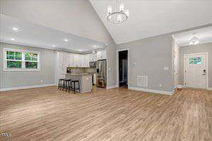 Open concept room with wood-style floors, light gray walls, recessed lighting, a white kitchen with stainless steel appliances, island seating, and double windows. Entryway with white door and a small overhead light.