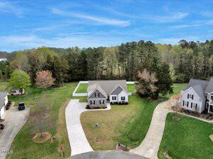 A single-story house with gray siding sits at the end of a curved driveway, surrounded by grass, trees, and neighboring homes. A large wooded area is visible behind the property under a blue sky.