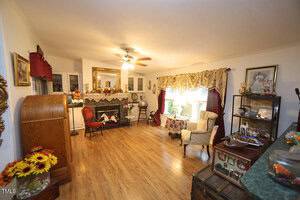 A living room with wooden floors, a ceiling fan, a fireplace with a mirror above it, several armchairs, decorative shelves, and large curtained windows letting in sunlight. Various knickknacks and flowers decorate the space.
