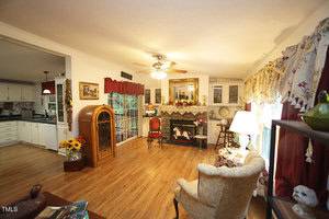 A cozy living room with wood floors, vintage furniture, a decorated fireplace, and floral curtains. The adjacent kitchen is visible through an open doorway on the left. A ceiling fan and lamp provide lighting.