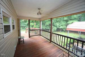 Covered wooden porch with ceiling fan, black railing, tan siding, and glass side table overlooks a green wooded backyard and a red-roofed carport. Steps lead down to a lower deck area.